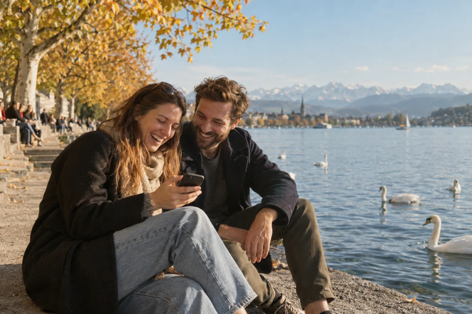 Deux amis assis au bord du lac de Zurich, en automne, rient devant un téléphone